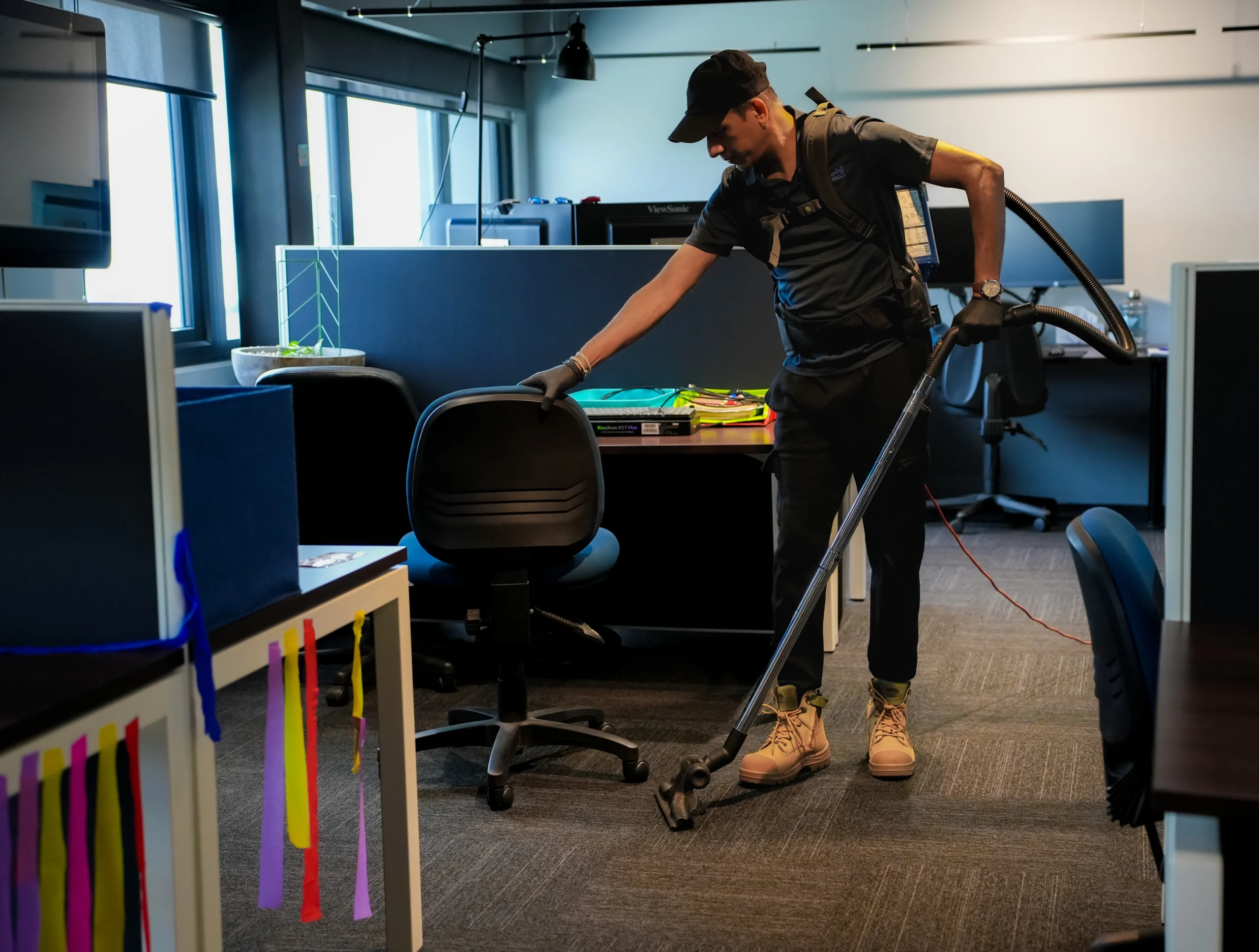 A Hobart Cleaning Company professional providing thorough janitorial services, using a backpack vacuum to clean the carpets around desks and workstations in a commercial office.